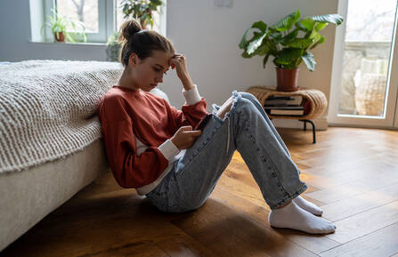Upset teen girl sitting on floor near bed using smartphone at home, scrolling social media