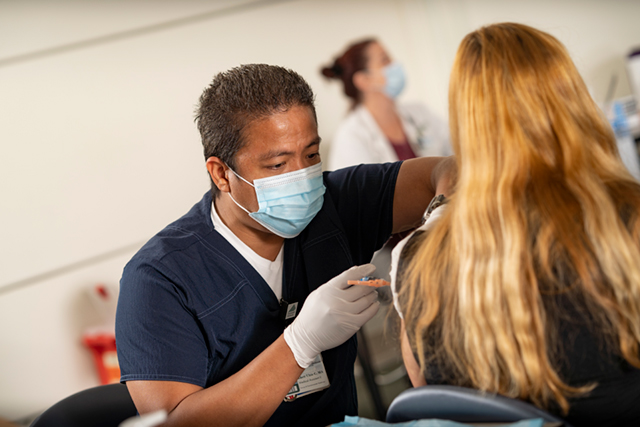 A healthcare worker in a blue uniform and mask is administering a vaccine to a patient.