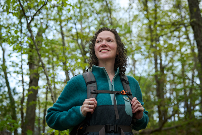 A woman with curly hair smiles while standing in a forest. She is wearing a teal fleece jacket and a backpack, looking up towards the trees and greenery around her. The scene is bright and captures the beauty of nature.