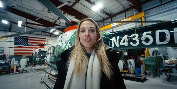 A young woman stands smiling in front of a helicopter ambulance in its hangar