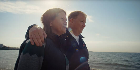 A man and woman walk arm in arm in front of a body of water on a sunny day
