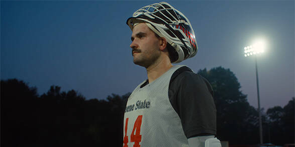 Young man in Lacrosse uniform stands with helmet raised, in front of stadium lighting at dusk