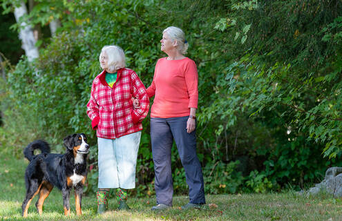 Two women, one elderly, stand by some greenery with a dog