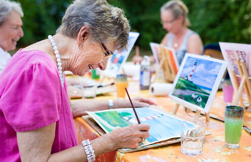 Senior woman painting a picture of a lighthouse at a table with other women