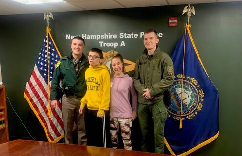 Two uniformed male New Hampshire State Troopers stand with two children, a boy on the left and a girl on the right, between them. They all stand between an American and New Hampshire state flag against a green wall.