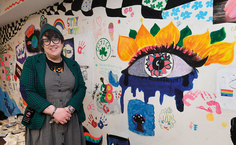  Art Therapist Mel Sack stands in front of a colorful mural on a wall, which features various artistic elements including an oversized eye with colorful flames, a rainbow flag, and playful doodles. The setting appears vibrant and creative.